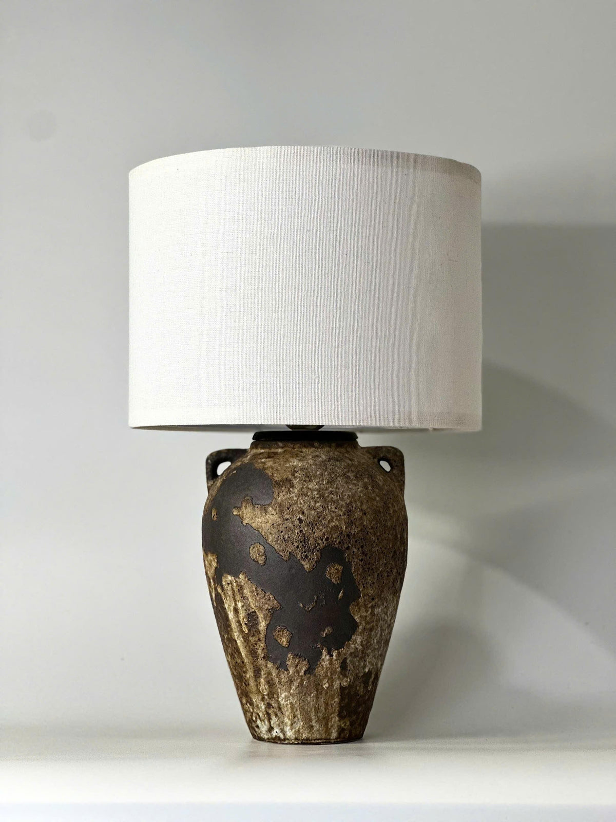Handmade ceramic table lamp with ancient side handles, a wabi-sabi silhouette, and soft fabric shade casting a warm, timeless glow.