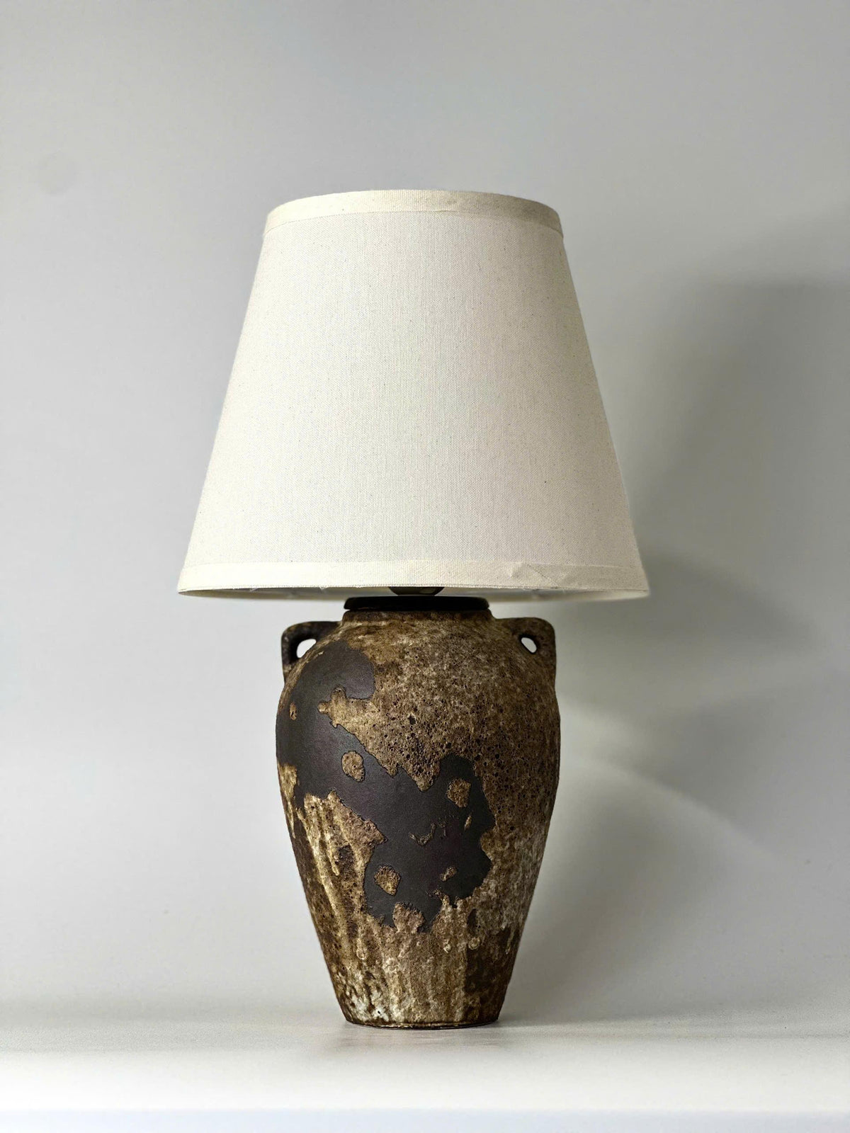 Handmade ceramic table lamp with ancient side handles, a wabi-sabi silhouette, and soft fabric shade casting a warm, timeless glow.