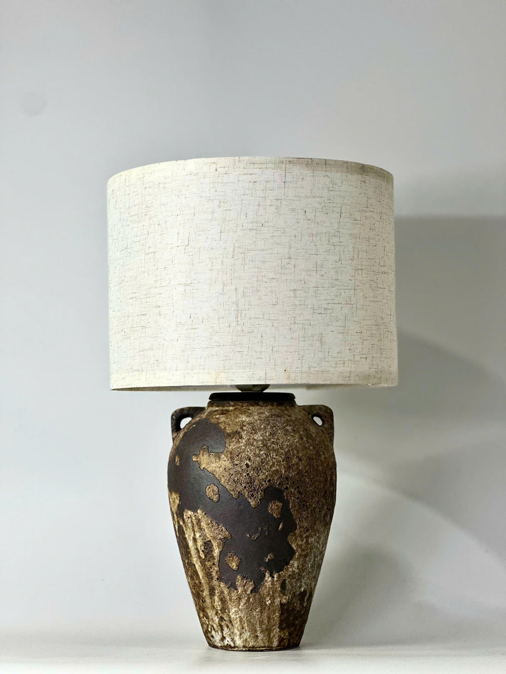 Handmade ceramic table lamp with ancient side handles, a wabi-sabi silhouette, and soft fabric shade casting a warm, timeless glow.