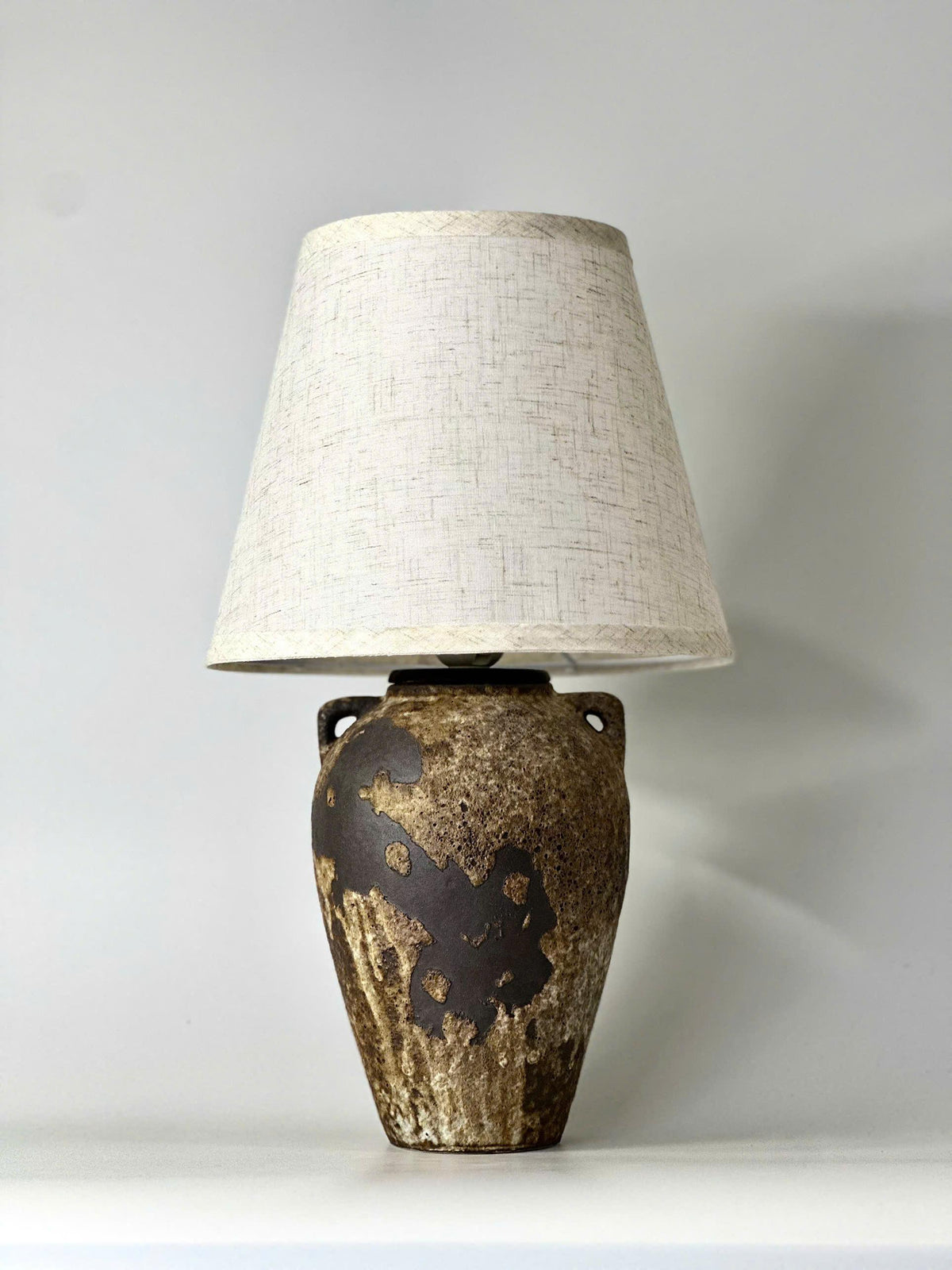 Handmade ceramic table lamp with ancient side handles, a wabi-sabi silhouette, and soft fabric shade casting a warm, timeless glow.